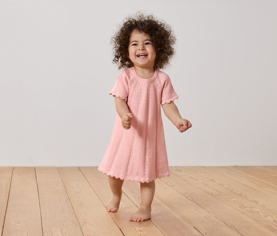 Un jeune enfant souriant aux cheveux bouclés portant une robe rose à ourlet ondulé sur un plancher de bois.