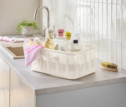 Un panier contenant des produits de nettoyage et un torchon rose est posé sur un comptoir de cuisine à côté d'un évier.