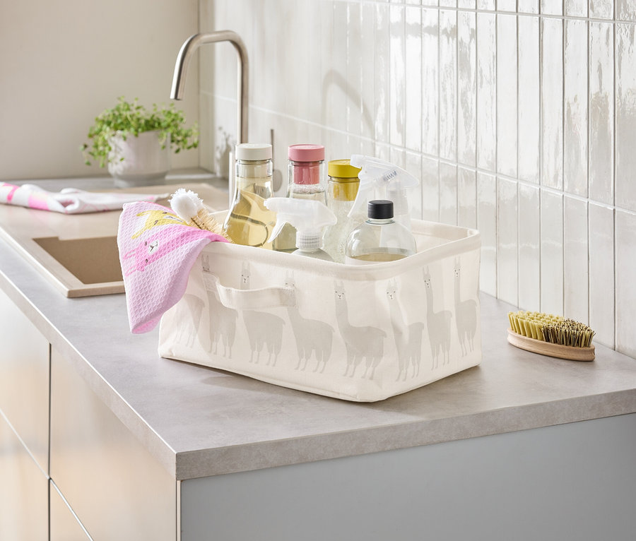 Un panier contenant des produits de nettoyage et un torchon rose est posé sur un comptoir de cuisine à côté d'un évier.