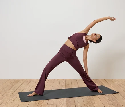 Une femme en tenue de sport bordeaux s'étire sur un tapis de yoga sur un plancher en bois devant un mur blanc.
