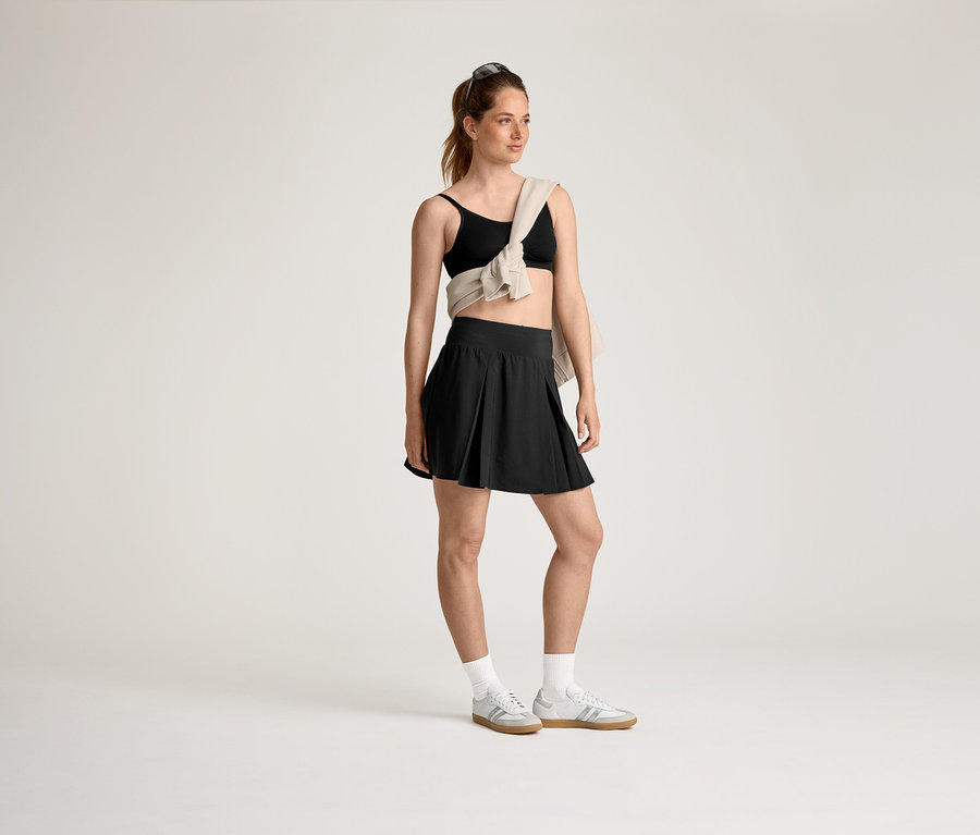 Femme portant un soutien-gorge de sport et une jupe noirs, un pull beige sur l'épaule et des baskets grises avec des chaussettes blanches.