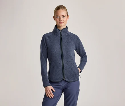 Femme aux cheveux blonds attachés, portant une veste polaire texturée bleu foncé avec un col montant et une fermeture éclair noire, ainsi qu'un pantalon cargo bleu foncé. Sa main gauche est dans une poche de la veste.