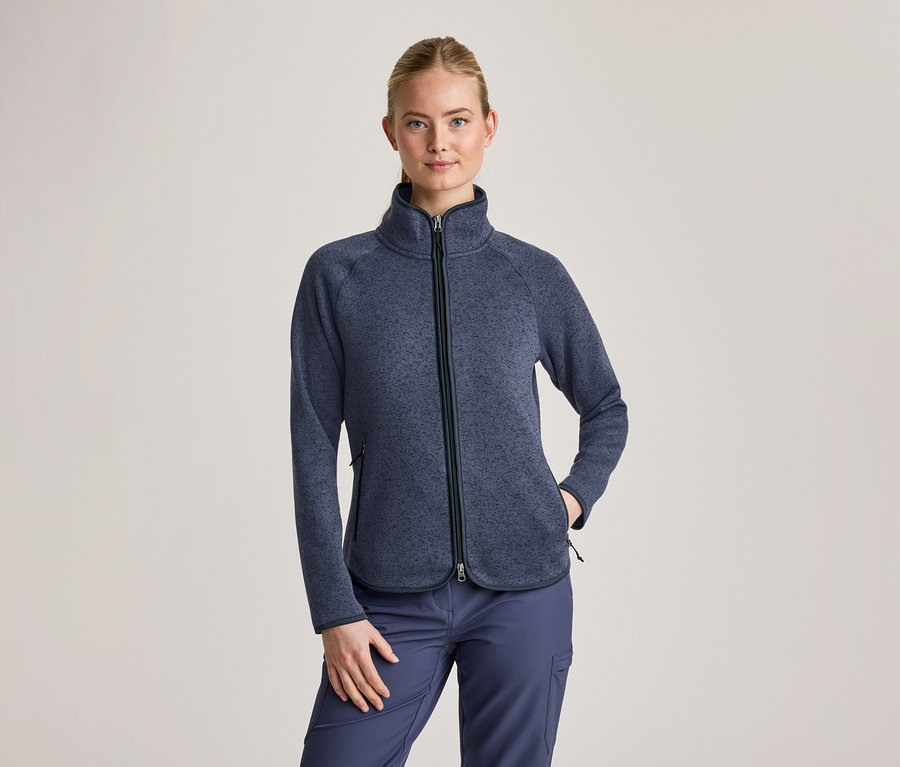 Femme aux cheveux blonds attachés, portant une veste polaire texturée bleu foncé avec un col montant et une fermeture éclair noire, ainsi qu'un pantalon cargo bleu foncé. Sa main gauche est dans une poche de la veste.