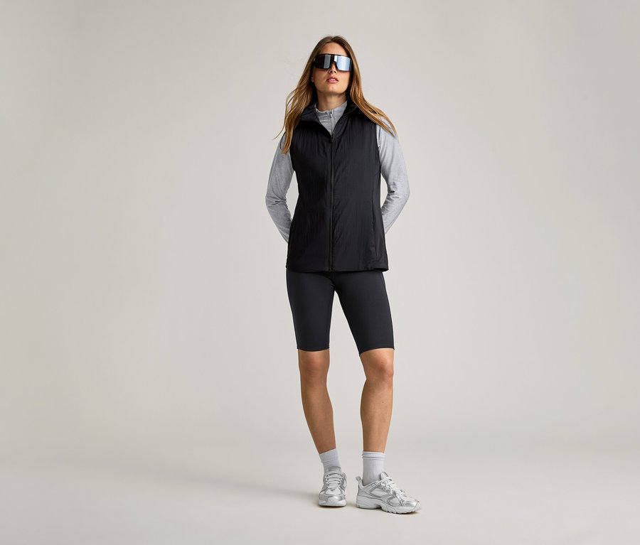 Femme portant des lunettes de soleil, un t-shirt gris, un cycliste noir, des chaussures de sport et un gilet de course noir.