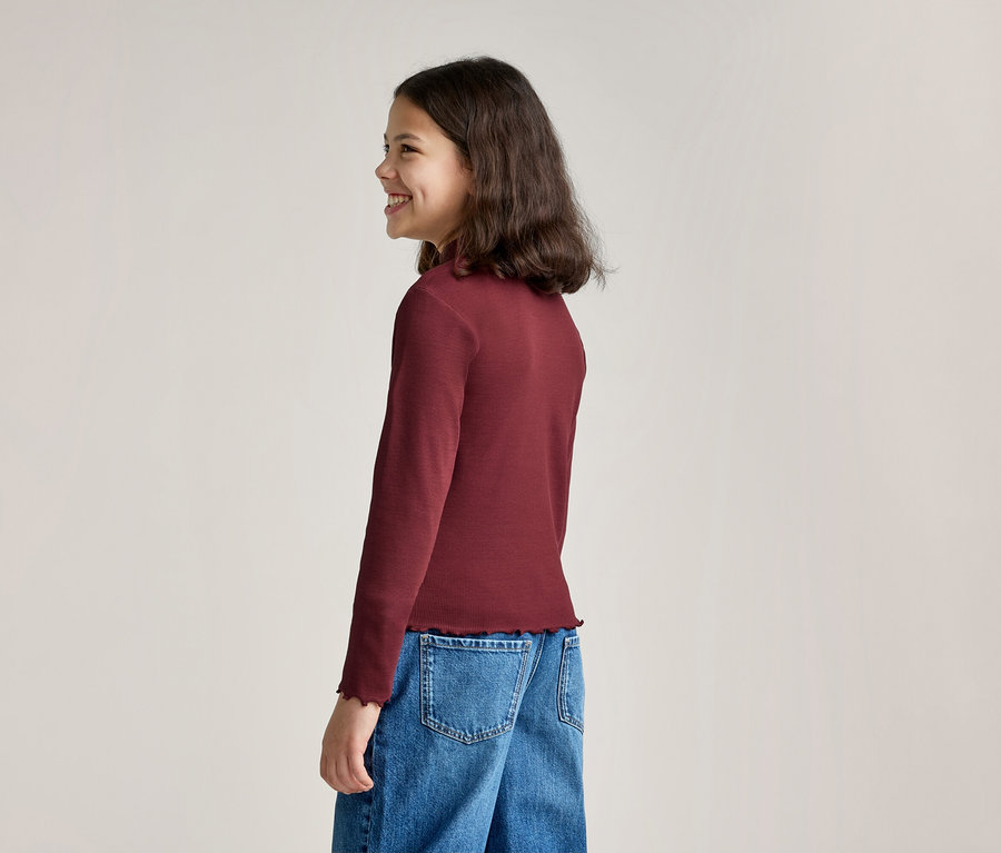 Une fille souriante aux cheveux longs et foncés est vue de profil, portant un haut bordeaux à manches longues avec un ourlet volanté et un jean bleu.