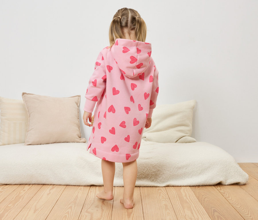 Une petite fille, dos à la caméra, porte une robe rose à capuche avec des motifs de cœurs.