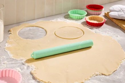 Sur un plan de travail fariné, une pâte étalée avec deux cercles découpés, un rouleau à pâtisserie vert et des moules à tartelettes colorés.