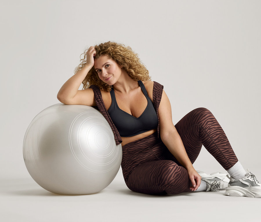 Femme aux cheveux bouclés assise à côté d'un ballon de gymnastique argenté, portant un soutien-gorge de sport noir, un legging à motif zébré et des baskets.