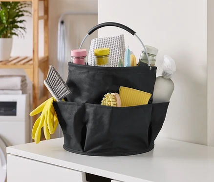 Photo d'un panier de nettoyage noir rempli de produits de nettoyage, incluant des brosses, des sprays et des chiffons, sur une surface blanche.