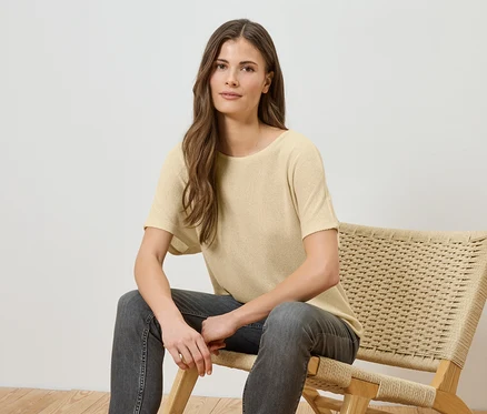 Une femme aux longs cheveux bruns est assise sur une chaise en osier, portant un t-shirt jaune clair et un jean gris.