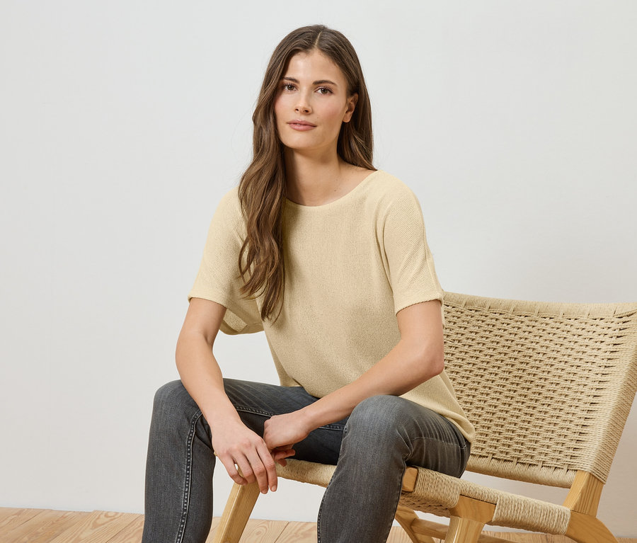 Une femme aux longs cheveux bruns est assise sur une chaise en osier, portant un t-shirt jaune clair et un jean gris.