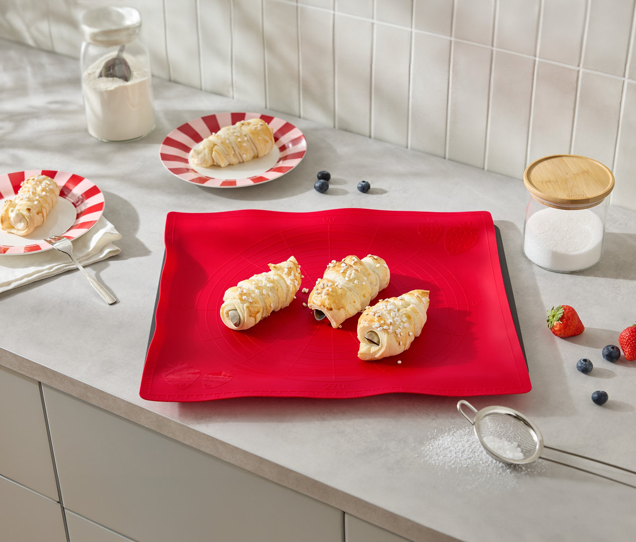 Trois croissants fourrés à la crème sont posés sur un tapis rouge, entourés d'ingrédients tels que des fraises et des myrtilles.