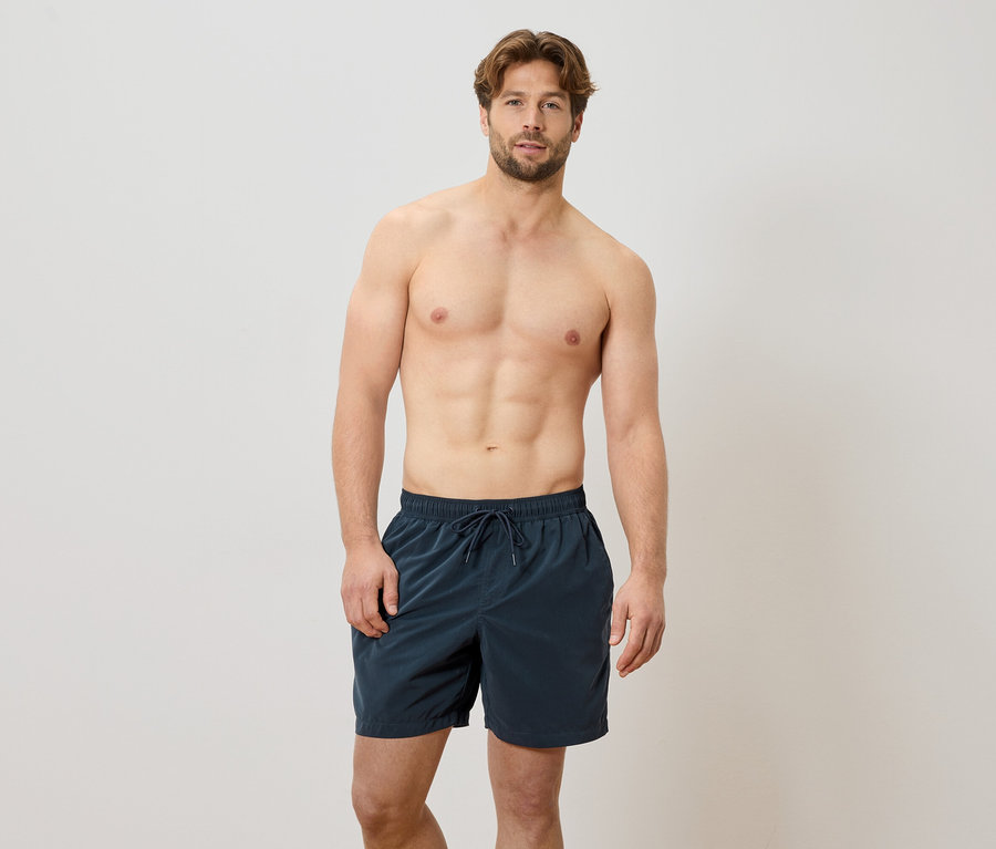 Homme debout pieds nus devant un mur blanc portant un short de bain bleu marine.