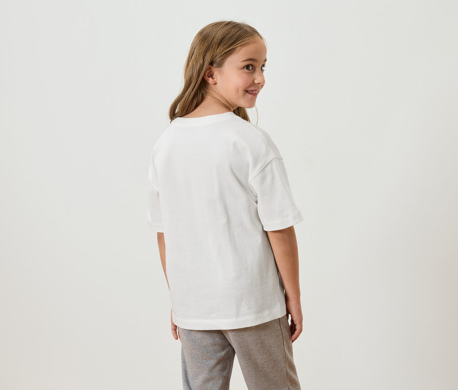 Une jeune fille aux longs cheveux bruns regarde par-dessus son épaule. Elle porte un t-shirt blanc et un pantalon de survêtement gris.