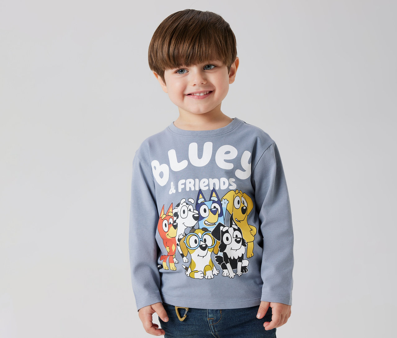 Un garçon souriant porte un t-shirt à manches longues pour enfant NAME IT « BLUEY » avec un motif Bluey et ses amis.