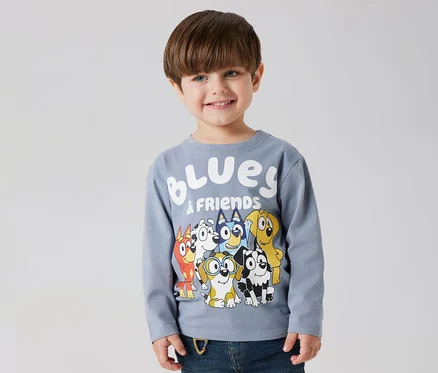 Un garçon souriant porte un t-shirt à manches longues pour enfant NAME IT « BLUEY » avec un motif Bluey et ses amis.