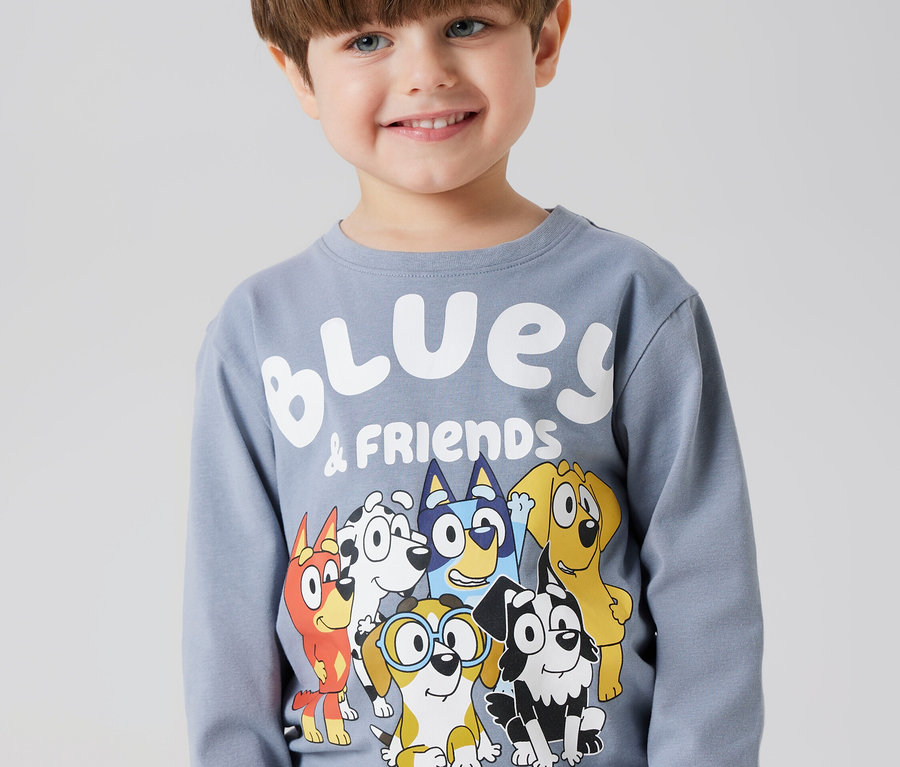 Un garçon souriant porte un T-shirt à manches longues gris avec une illustration de Bluey et ses amis.