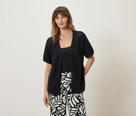 Une femme porte un cardigan en maille noir à manches courtes sur un haut noir et un pantalon à motifs noirs et blancs.