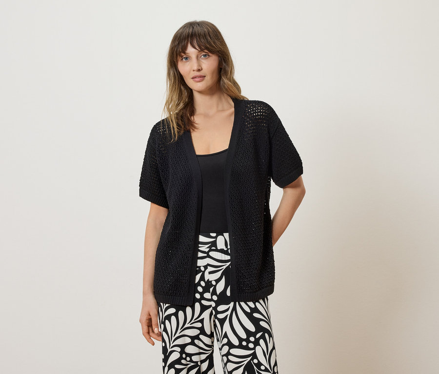 Une femme porte un cardigan en maille noir à manches courtes sur un haut noir et un pantalon à motifs noirs et blancs.