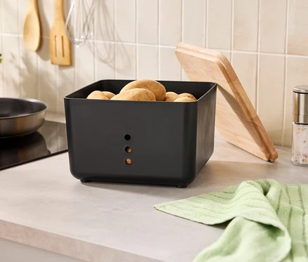 Sur un comptoir de cuisine se trouvent un récipient à pommes de terre noir rempli de pommes de terre, une planche à découper, des moulins à épices et un torchon.