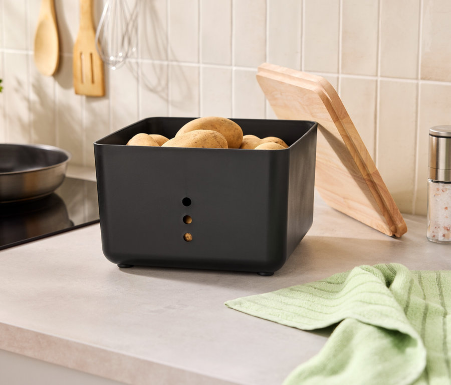 Sur un comptoir de cuisine se trouvent un récipient à pommes de terre noir rempli de pommes de terre, une planche à découper, des moulins à épices et un torchon.