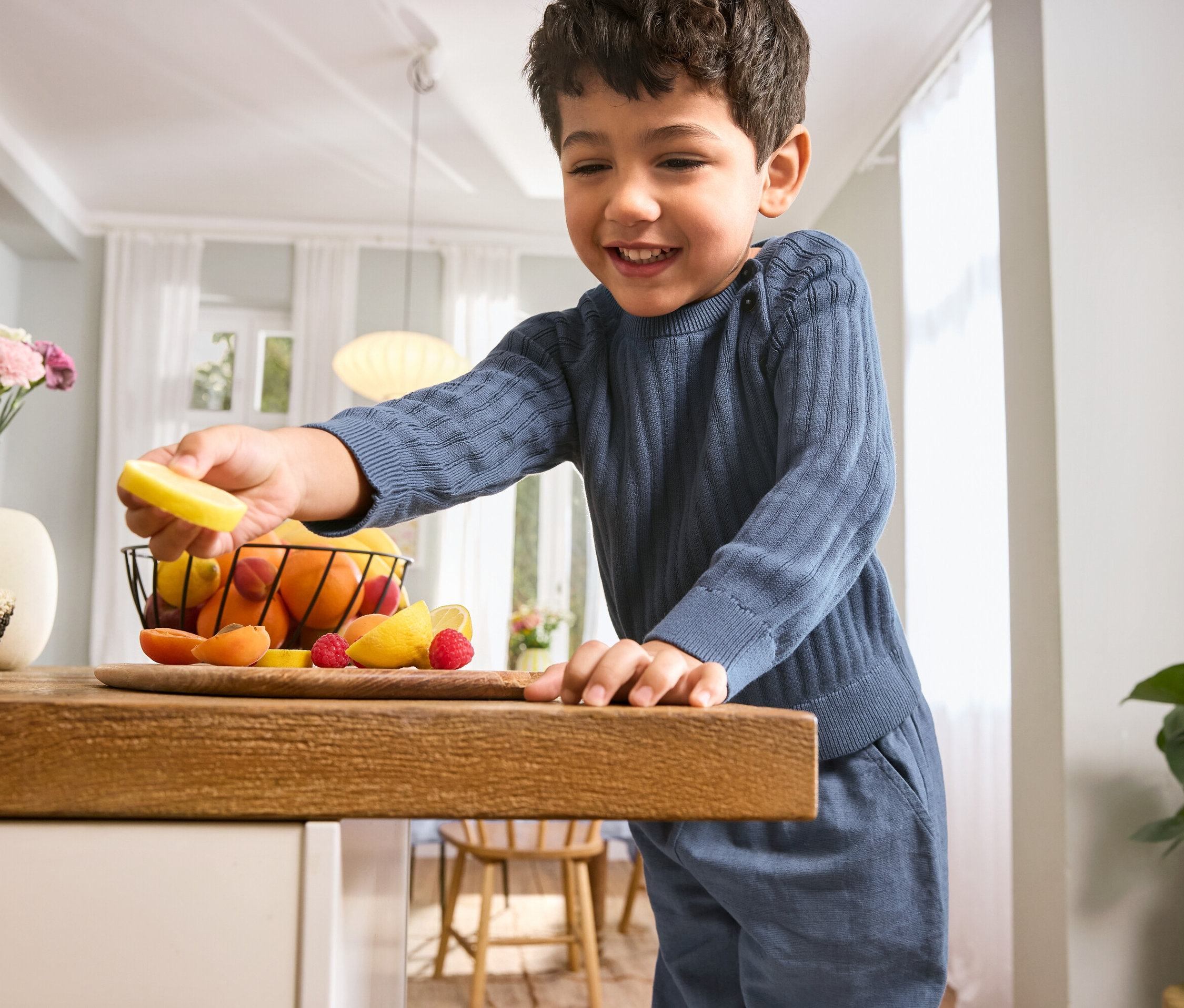 Un garçon portant un pull en tricot pour enfants et un pantalon à enfiler pour enfants tient une tranche de citron au-dessus d'une table avec des fruits.