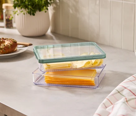Sur un comptoir de cuisine, deux récipients transparents avec des tranches de fromage, un bagel, un bol de guacamole et des épices.