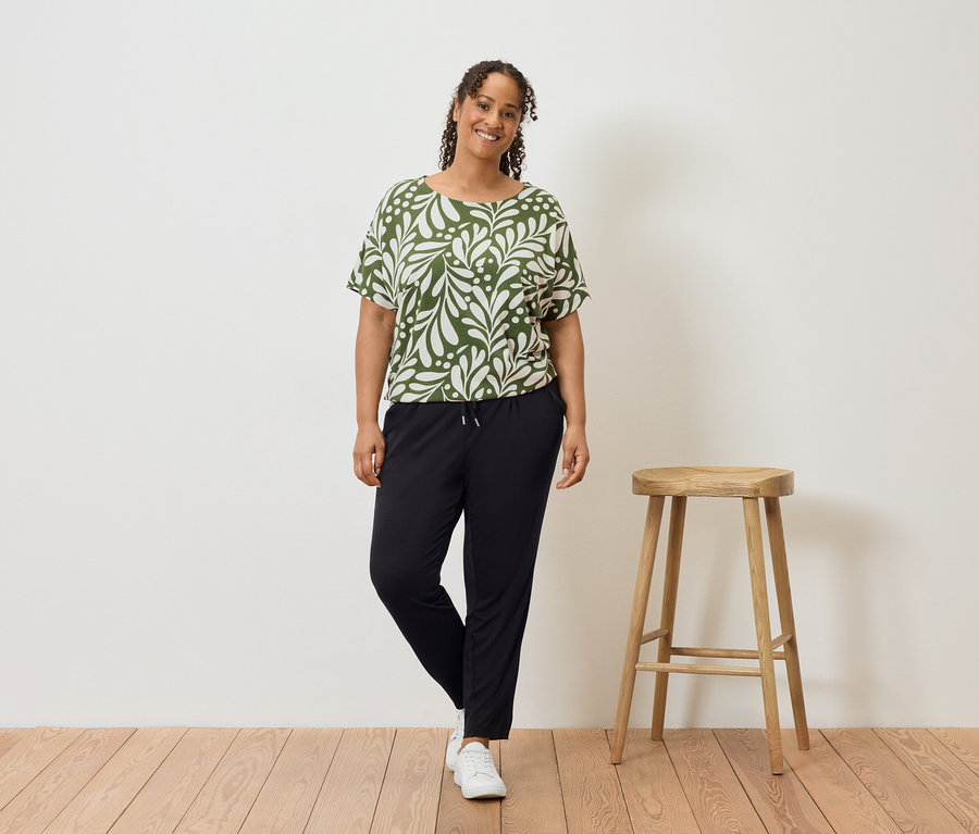 Une femme aux cheveux bouclés se tient à côté d'un tabouret, portant un haut vert et blanc, un pantalon noir et des baskets blanches.