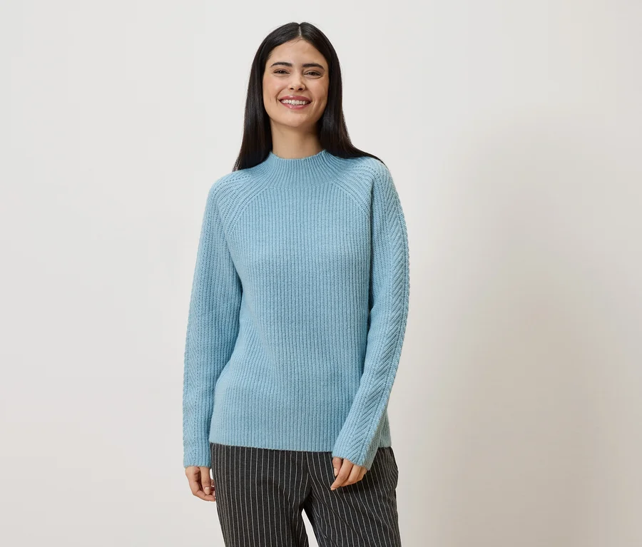 Une femme aux longs cheveux foncés porte un pull bleu clair et un pantalon rayé sur un fond blanc.