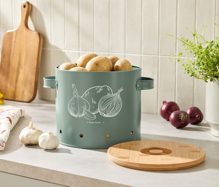 Pommes de terre dans une boîte décorée d'oignons et d'ail. À côté, des gousses d'ail, des oignons rouges, une planche à découper, une bouteille d'huile et une plante en pot.