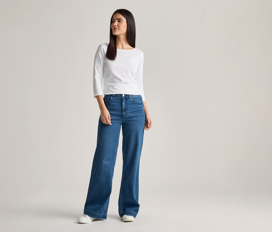 Une femme aux longs cheveux foncés porte un chemisier blanc à manches longues, un jean bleu large et des chaussures blanches sur un fond clair.
