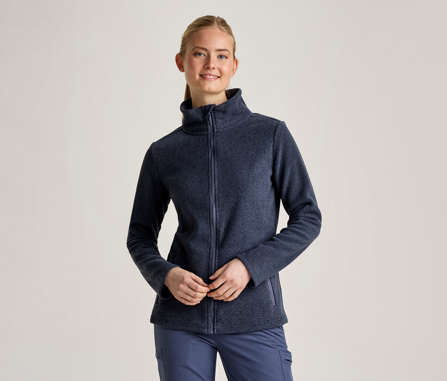 Une femme souriante aux cheveux blonds attachés porte une veste polaire zippée bleu foncé et un pantalon bleu, les mains jointes devant elle.