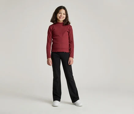 Une jeune fille souriante aux cheveux bruns mi-longs porte un haut à manches longues bordeaux et un pantalon noir évasé.