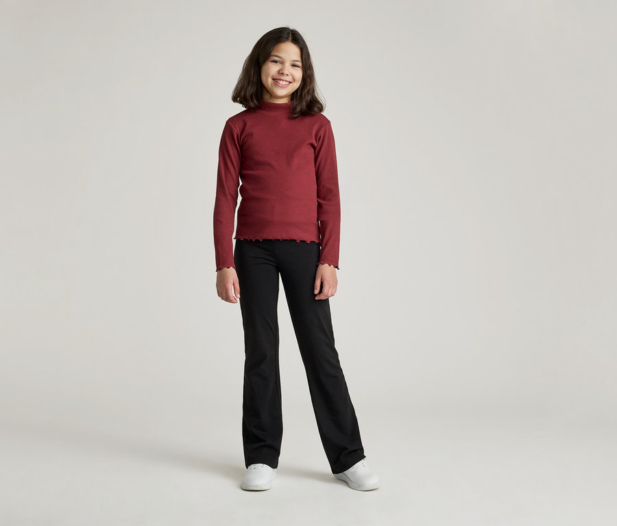 Une jeune fille souriante aux cheveux bruns mi-longs porte un haut à manches longues bordeaux et un pantalon noir évasé.