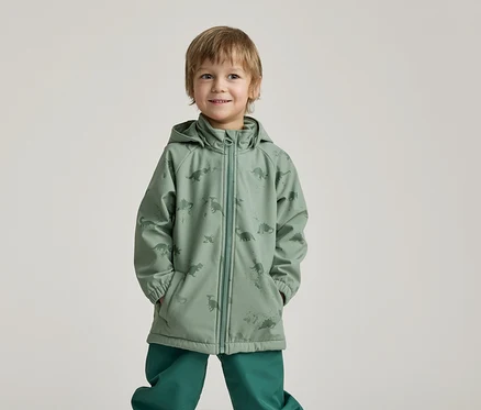Un garçon souriant, les mains dans les poches, porte un imperméable vert à motifs de dinosaures et un pantalon vert sur un fond clair.
