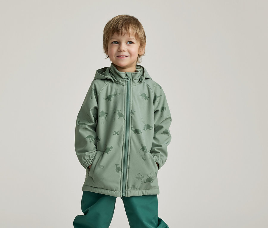 Un garçon souriant, les mains dans les poches, porte un imperméable vert à motifs de dinosaures et un pantalon vert sur un fond clair.