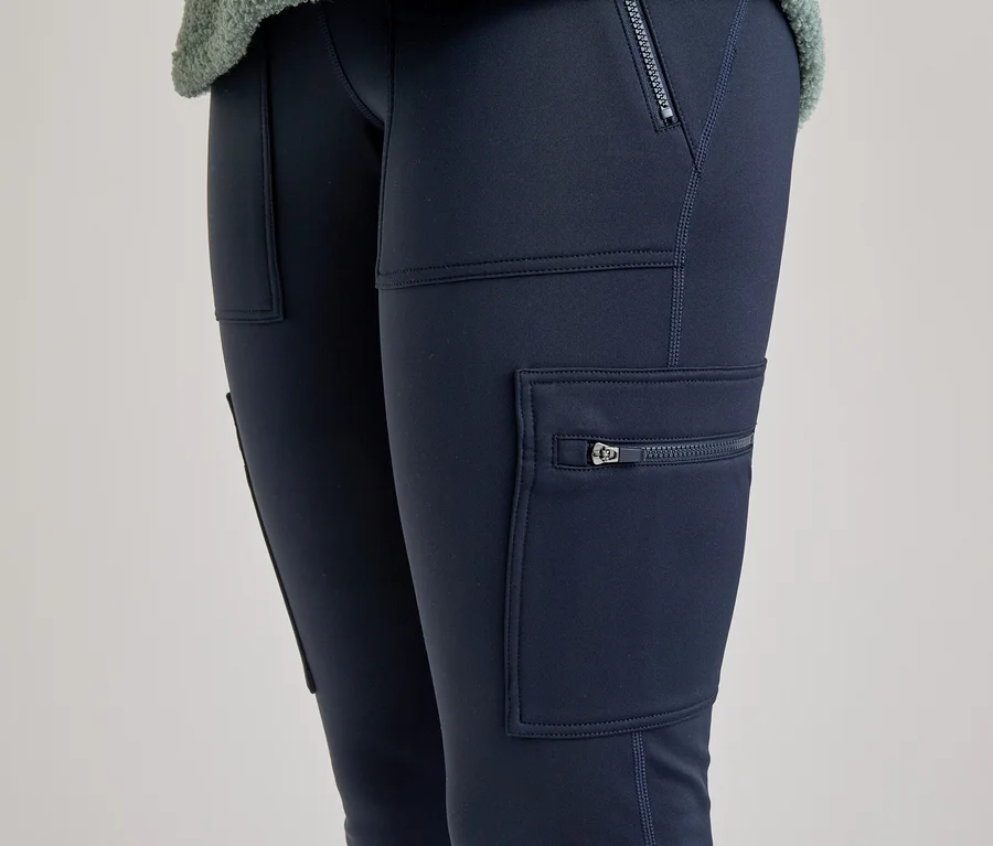 Gros plan sur les jambes d'une personne portant un legging cargo bleu foncé avec des poches zippées et des poches plaquées. Une partie d'un haut vert clair et duveteux est visible en haut.
