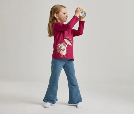 Une fillette portant un t-shirt rose avec un motif de raton laveur et un jean bleu pose avec un appareil photo jouet sur un fond clair.