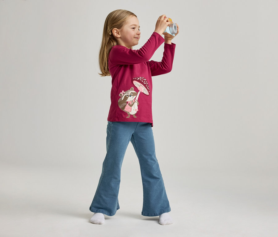 Une fillette portant un t-shirt rose avec un motif de raton laveur et un jean bleu pose avec un appareil photo jouet sur un fond clair.