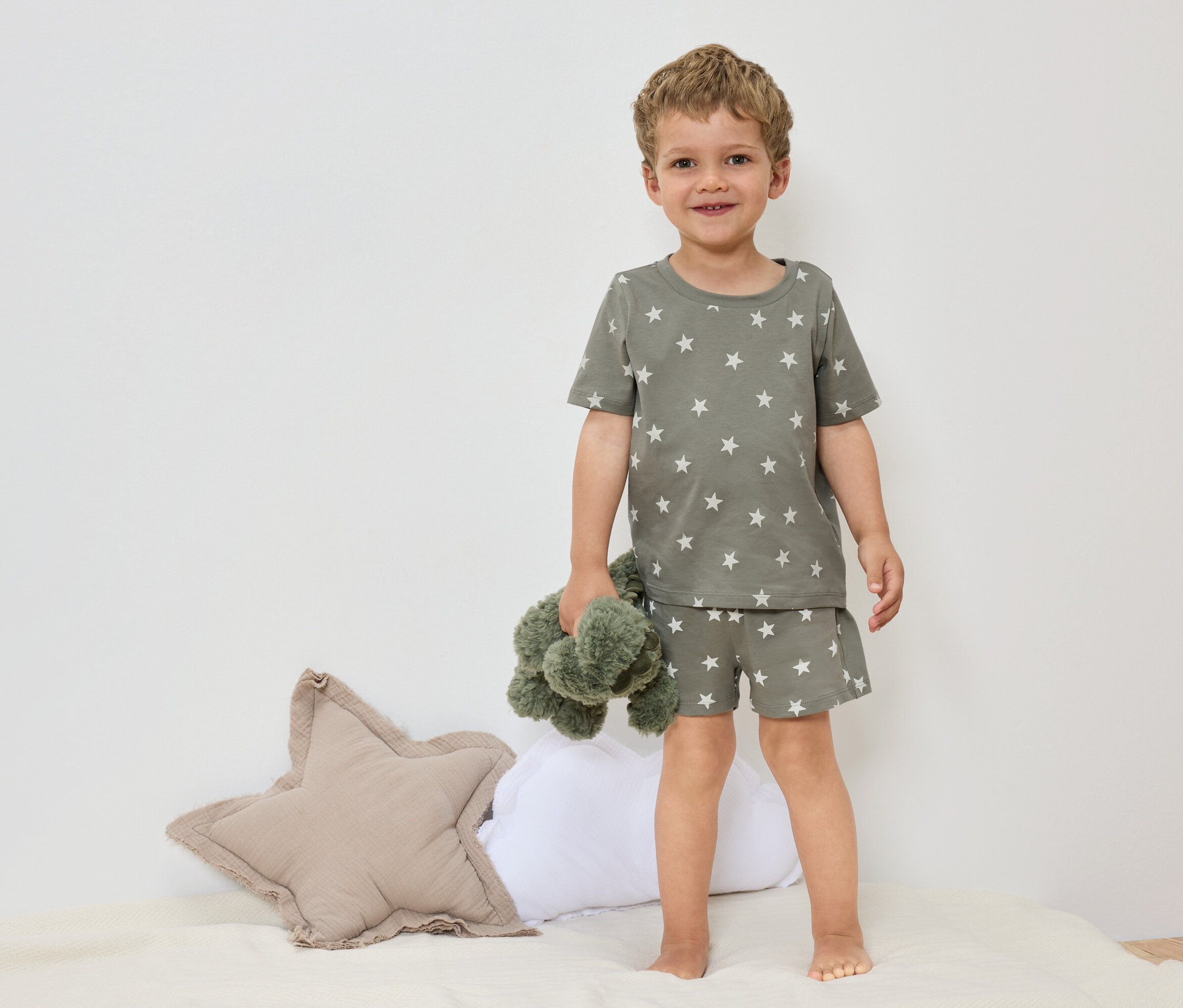 Un petit garçon se tient pieds nus sur un lit, vêtu d'un pyjama vert olive à motifs d'étoiles et tenant une peluche verte.