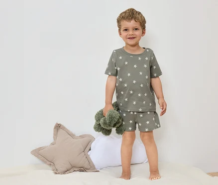 Un petit garçon se tient pieds nus sur un lit, vêtu d'un pyjama vert olive à motifs d'étoiles et tenant une peluche verte.