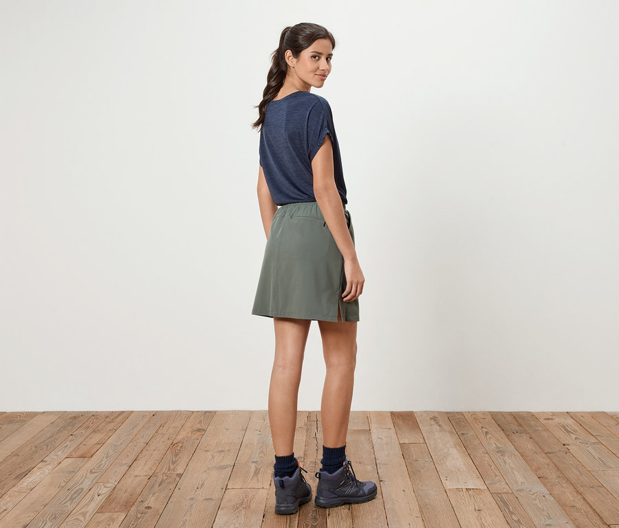 Une femme se tient sur un plancher en bois, portant un t-shirt bleu et une jupe verte.