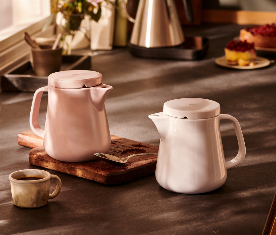 Deux cafetières en céramique, blanche et rose, sont posées côte à côte sur une planche en bois.