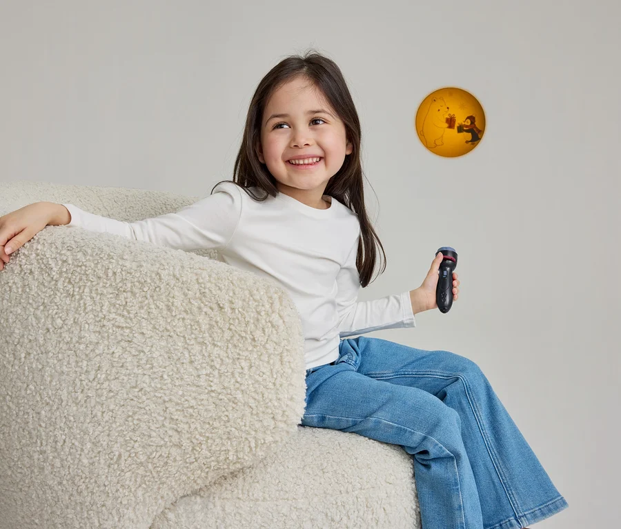 Une jeune fille souriante aux longs cheveux foncés est assise dans un fauteuil moelleux de couleur crème. Elle porte un t-shirt blanc à manches longues et un jean bleu, tient un projecteur jouet noir dans sa main droite et regarde une projection circulaire jaune-orange sur le mur, représentant un ours polaire et une personne échangeant des cadeaux.