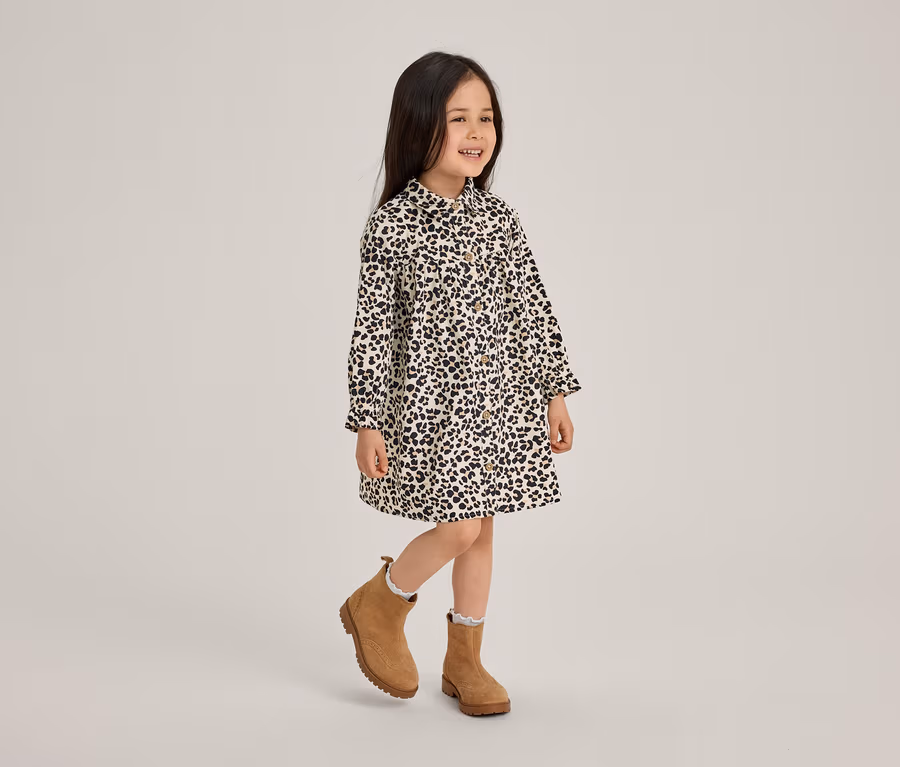 Une petite fille souriante vêtue d'une robe à imprimé léopard noir et blanc et de bottines marron se tient sur une jambe.