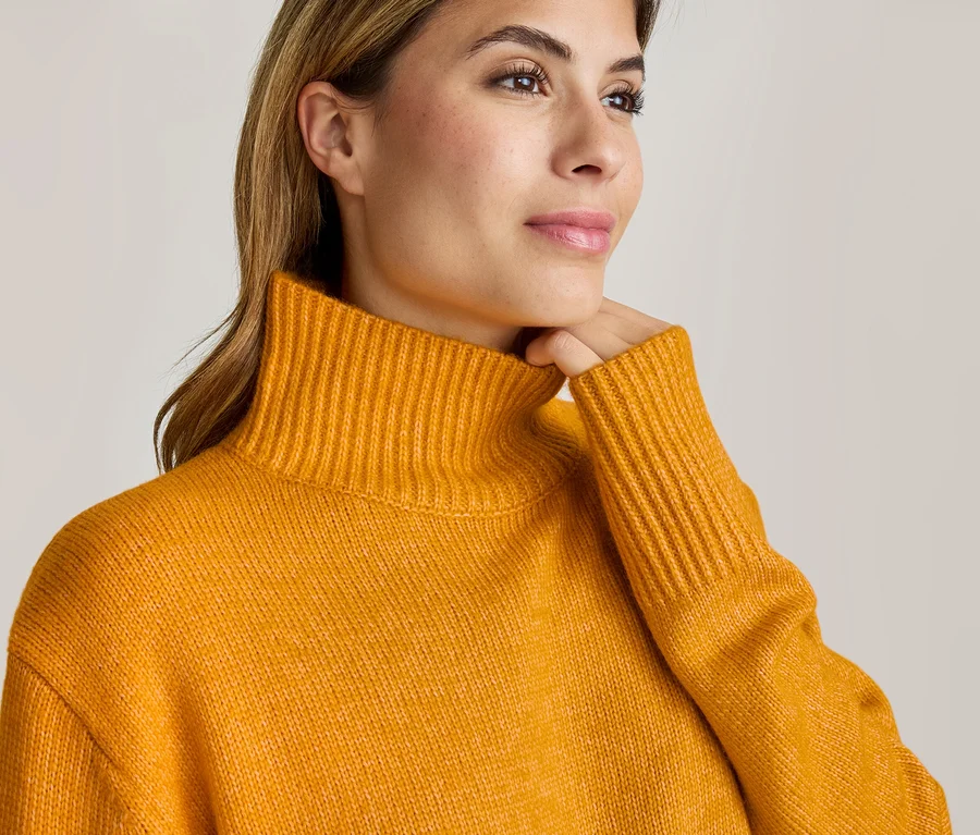 Gros plan d'une femme portant un pull à col roulé jaune moutarde, regardant vers la droite et touchant délicatement son menton.