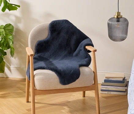Fauteuil beige avec un plaid en fausse fourrure bleu foncé, à côté d'une grande plante Monstera dans un panier tressé. Une pile de livres est au sol et une lampe en verre côtelé est suspendue au-dessus. Une literie à motifs bleu clair est visible à droite.