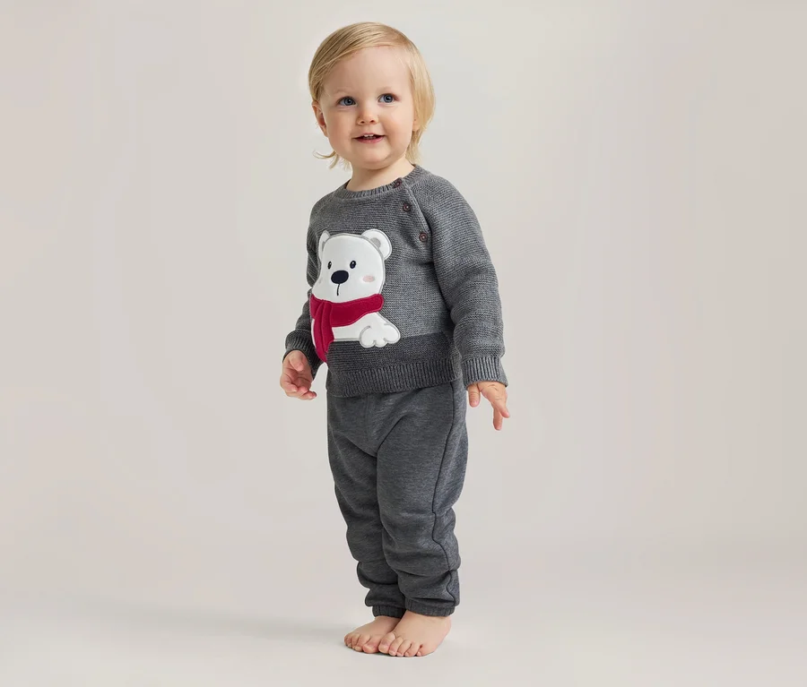 Bébé debout, souriant, portant un pull gris en tricot avec un ours polaire blanc à écharpe rouge et un pantalon gris, pieds nus.