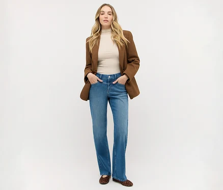 Femme aux longs cheveux blonds debout, portant un blazer marron sur un haut beige, un jean bleu et des ballerines imprimé léopard, les mains dans les poches.