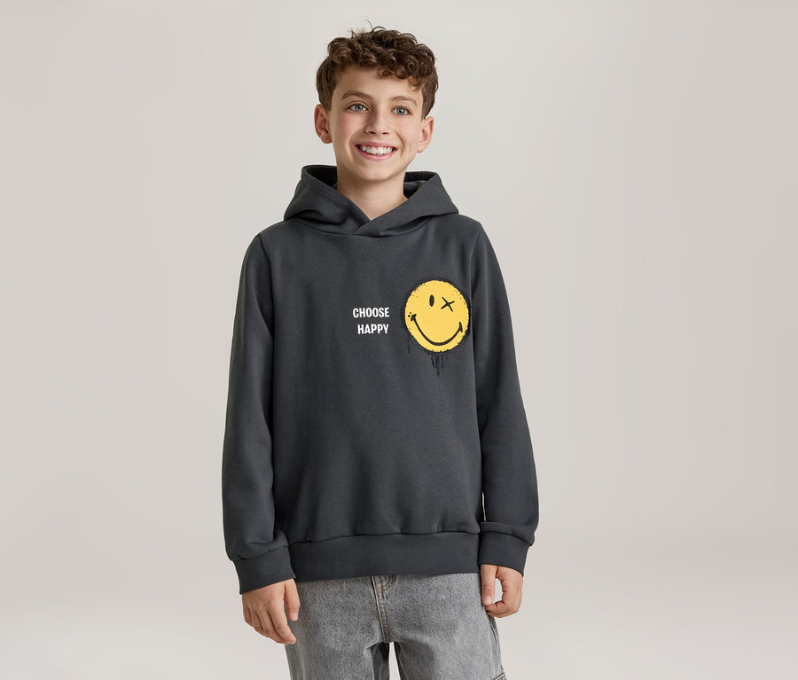 Jeune garçon souriant portant un sweat à capuche gris foncé avec un smiley jaune dont un œil est un X et qui a un effet de fonte, et l'inscription « CHOOSE HAPPY ». Il porte un jean gris clair.